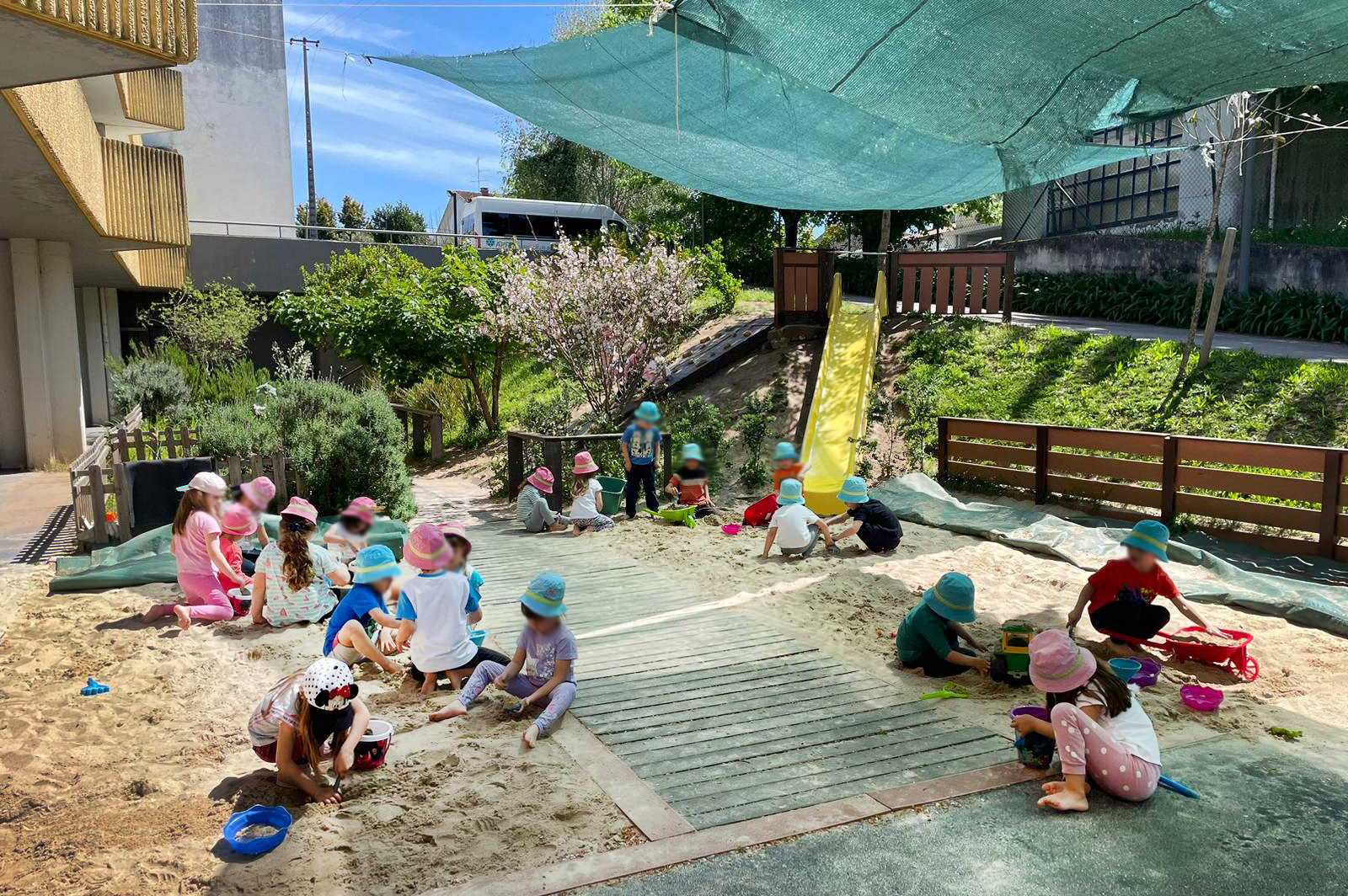 Perto de duas dezenas de crianças brincam em espaço exterior com areia. No meio um passadiço de madeira e ao fundo vê-se um jardim primaveril. Rosto das crianças desfocados.