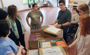 Oito pessoas em semí-círculo junto de uma mesa com seis caixas contendo várias dezenas de livros.