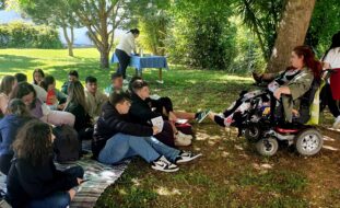 Fotografia de jardim exterior da Villa Urbana de Valbom. Na relva, e em cima de uma manta colorida, estão 14 jovens. Ao fundo uma mesa com uma pessoa a preparar algo. à direita da imagem, sentada em cadeira de rodas, pessoa adulta que conversa com os jovens.