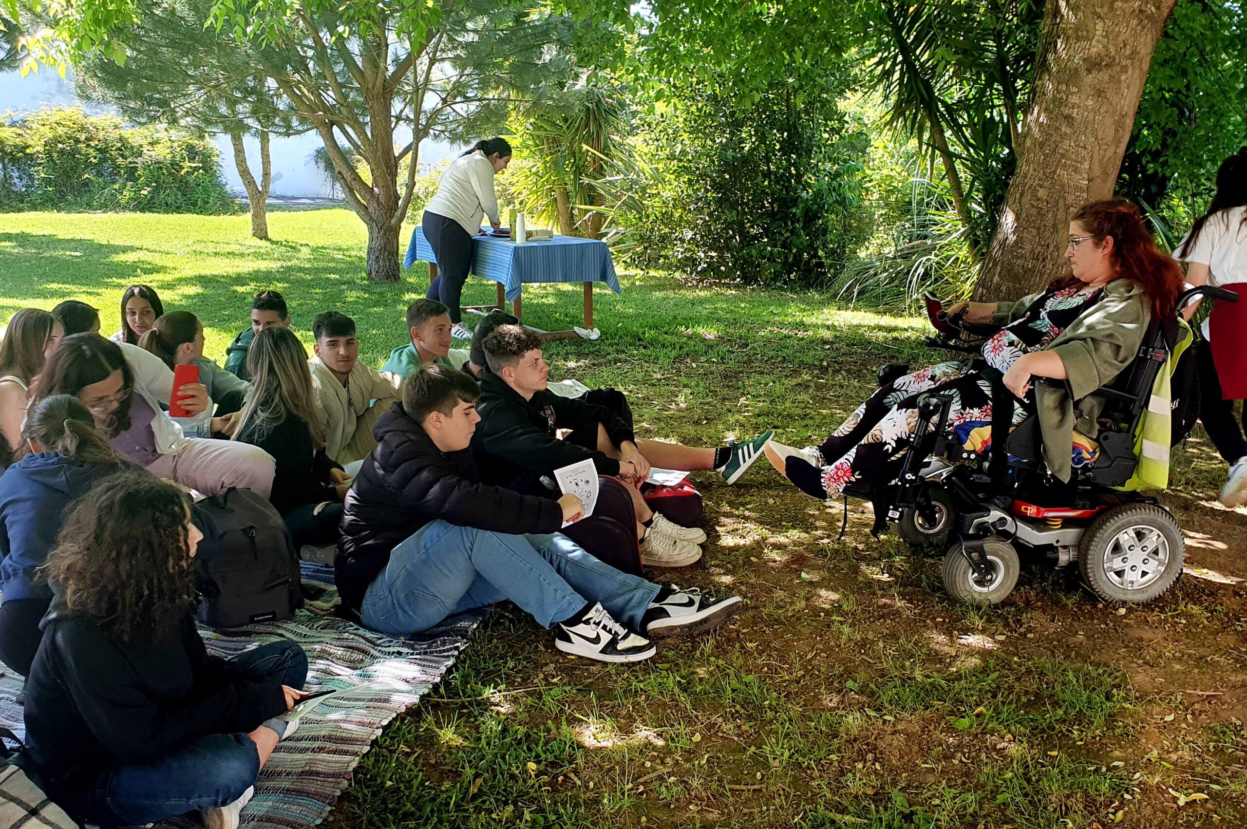 Fotografia de jardim exterior da Villa Urbana de Valbom. Na relva, e em cima de uma manta colorida, estão 14 jovens. Ao fundo uma mesa com uma pessoa a preparar algo. à direita da imagem, sentada em cadeira de rodas, pessoa adulta que conversa com os jovens.