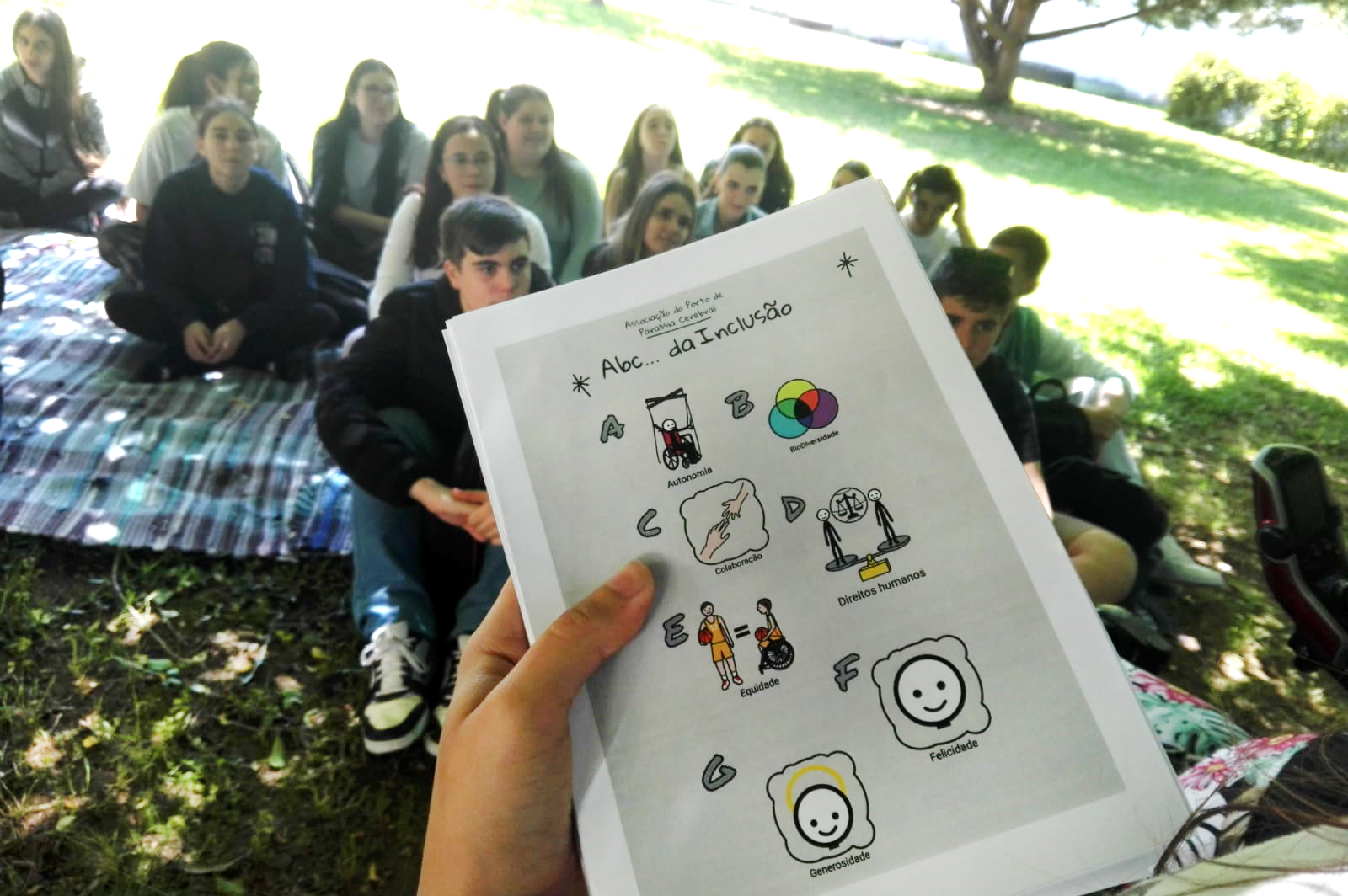 Grande plano de uma mão a segurar alguns exemplares do manual ABC da Inclusão. Mais ao fundo, ligeiramente desfocados, jovens sentados em manta pousada na relva.