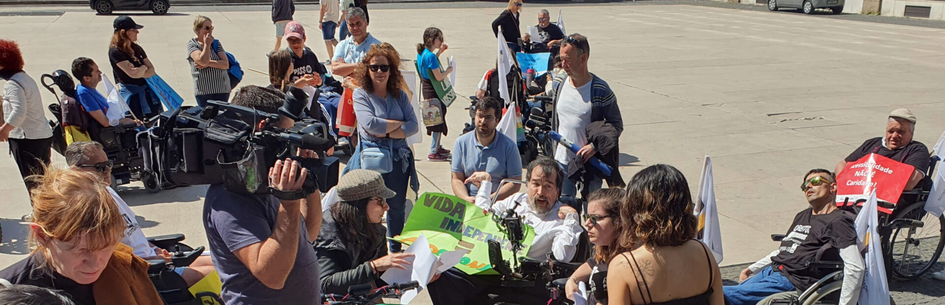 Grupo de pessoas em concentração na Praça D. João I, no Porto. Ao centro, em cadeira de rodas, alguns elementos da organização preparam-se para entrevista. Vê-se ainda repórter de imagem e jornalista de televisão.