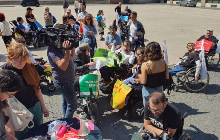 Grupo de pessoas em concentração na Praça D. João I, no Porto. Ao centro, em cadeira de rodas, alguns elementos da organização preparam-se para entrevista. Vê-se ainda repórter de imagem e jornalista de televisão.
