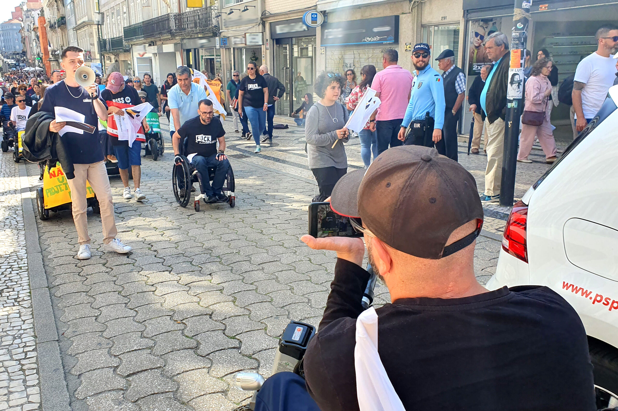 Manifestantes em marcha pela Rua de Santa Catarina, no Porto. À frente da comitiva segue uma pessoa com um megafone. Em primeiro plano está uma pessoa sentada em cadeira de rodas a filmar, com telemóvel, a manifestação. Várias pessoas assistem na zona lateral.