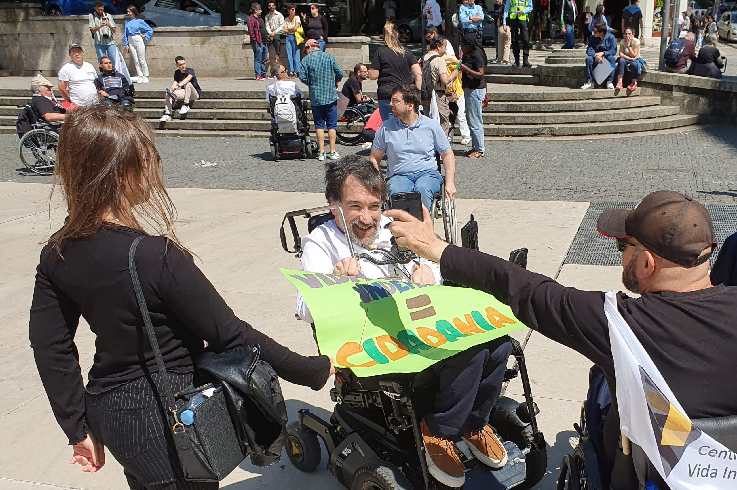 Grupo de pessoas em concentração na Praça D. João I, no Porto. Uma pessoa em cadeira de rodas sorri ao ver uma fotografia que lhe é mostrada num telemóvel.