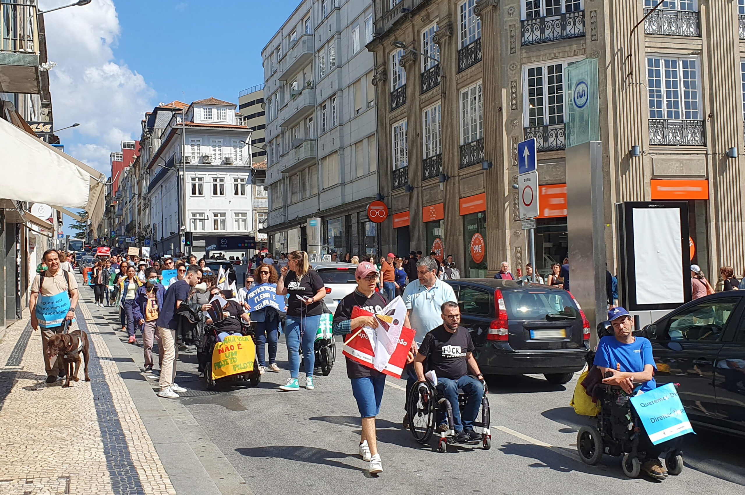 Manifestantes em marcha. Alguns seguram cartazes com reivindicações. À esquerda segue pessoa invisual com cão-guia.
