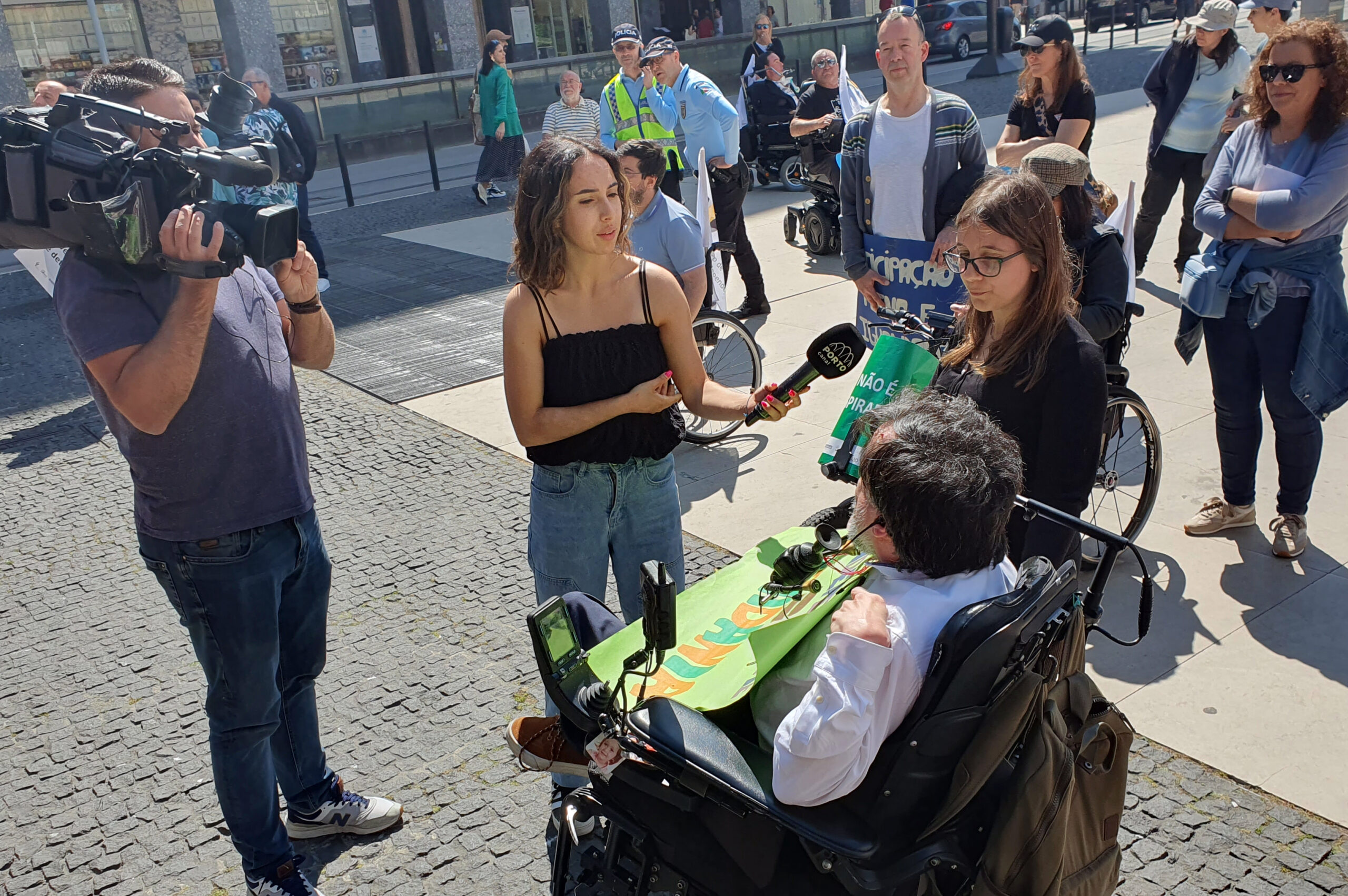 José Rui Marques, representante da APPC na Comissão Organizadora, a ser entrevistado pelo Porto Canal. O repórter de imagem filma o elemento da APPC e a jornalista recolhe depoimento junto da assistente pessoal. Algumas pessoas assistem e, ao fundo, estão dois elementos da PSP.