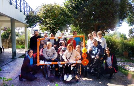 Fotografia de grupo com perto de duas dezenas de pessoas. À frente de algumas delas está uma moldura, laranja, com as expressões “Dia da Família” e “Casa da Maceda”. Ao fundo algumas árvores e parte do jardim da Casa da Maceda.
