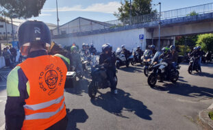 Em grande plano, de costas, participante no passeio com um colete laranja vestido. No colete a inscrição “Ride to share”, um rosto estilizado de homem com barba a usar capacete e óculos de sol. O referido rosto é enquadrado pela imagem de duas mãos. Ao fundo, alinhados, vários participantes e as respetivas motos.