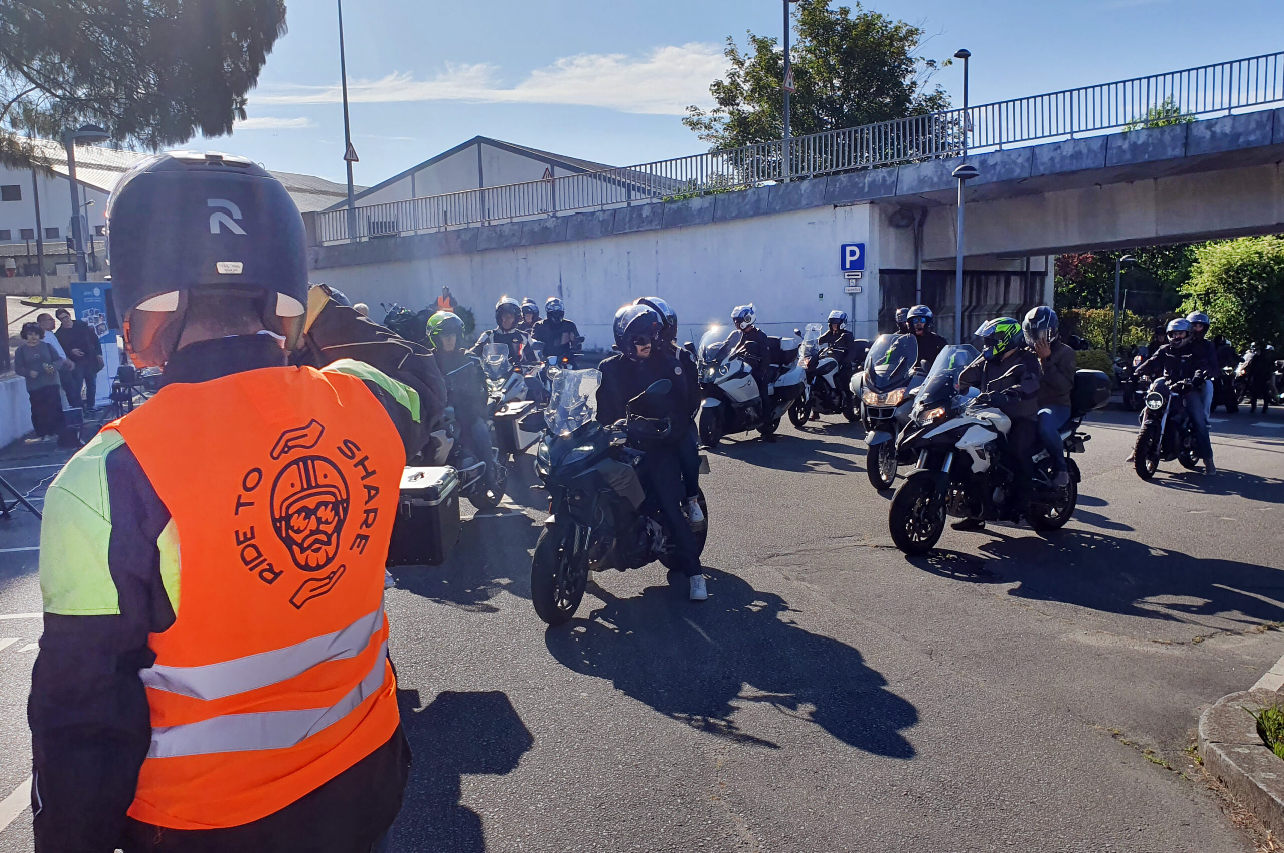 Em grande plano, de costas, participante no passeio com um colete laranja vestido. No colete a inscrição “Ride to share”, um rosto estilizado de homem com barba a usar capacete e óculos de sol. O referido rosto é enquadrado pela imagem de duas mãos. Ao fundo, alinhados, vários participantes e as respetivas motos.