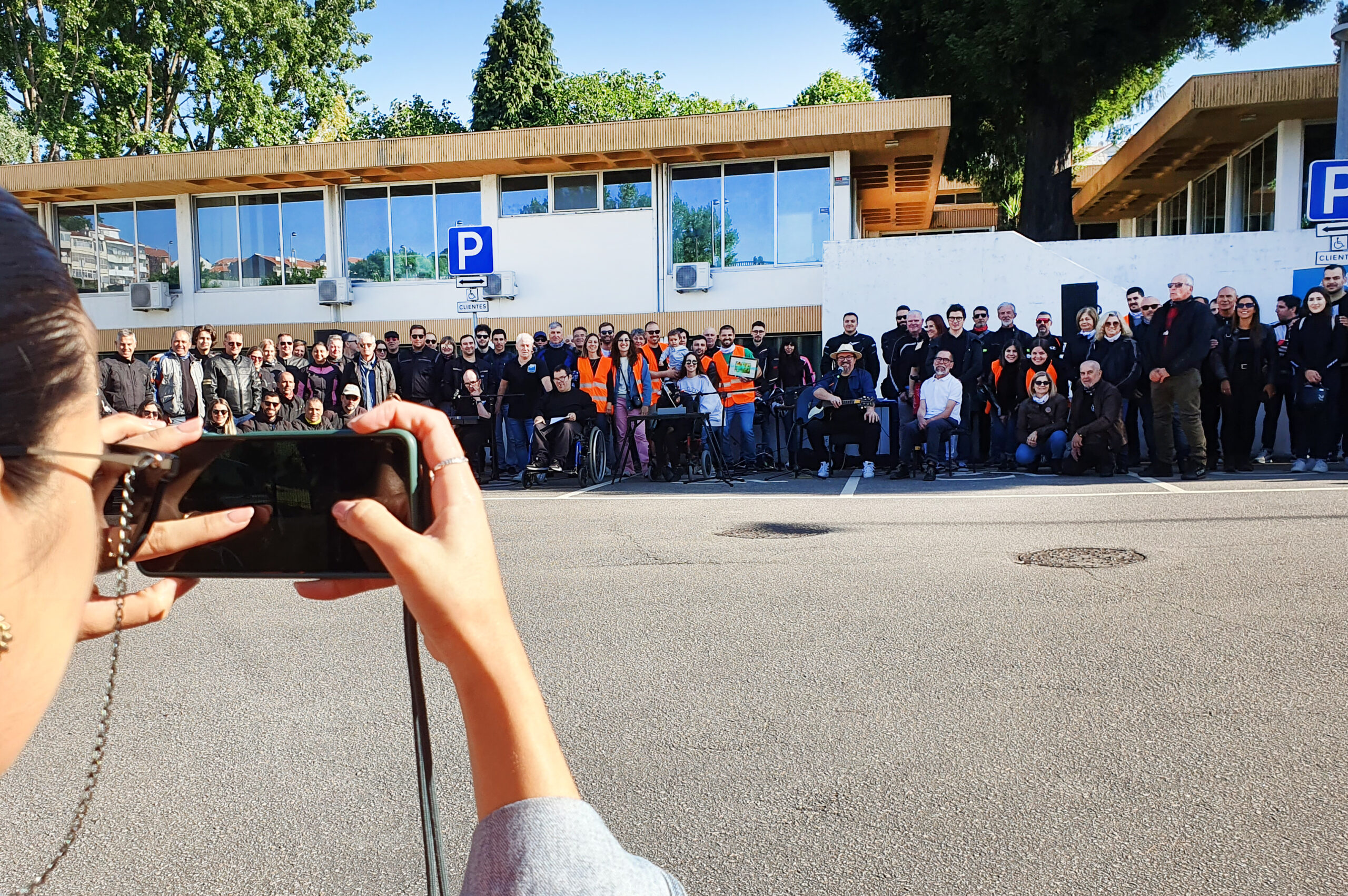 Ao fundo da imagem, alinhados, muitos dos participantes no passeio solidário. Vê-se ainda parte do Centro de Reabilitação. Em primeiro plano parte de um rosto e mãos a usar um telemóvel para fotografia de grupo.