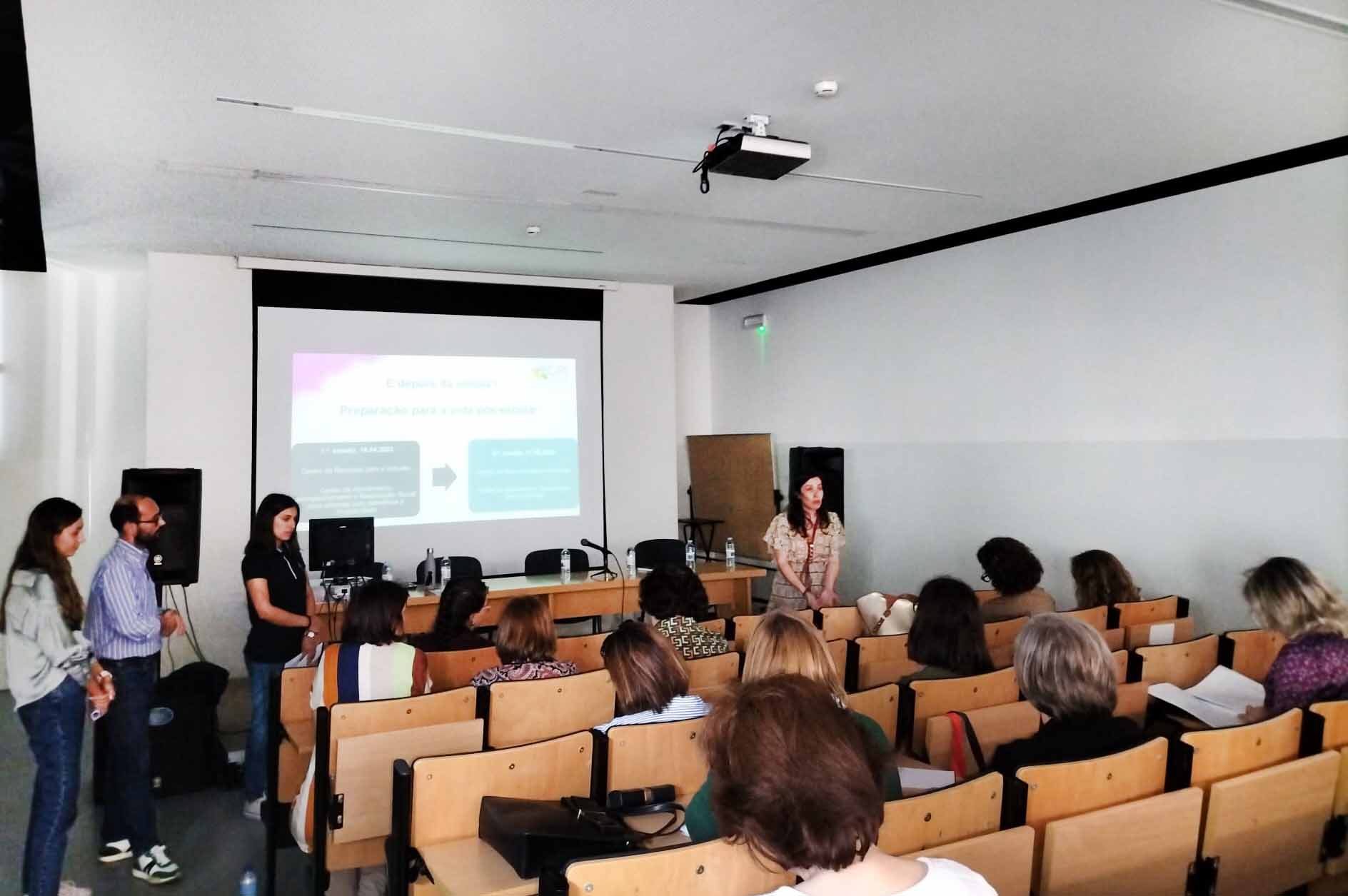 Fotografia de auditório de estabelecimento de ensino. Ao fundo, junto de quadro com projeção, mesa e cadeiras, estão quatro pessoas em pé. Em primeiro plano, de costas, assistem várias pessoas sentadas nas cadeiras do auditório.