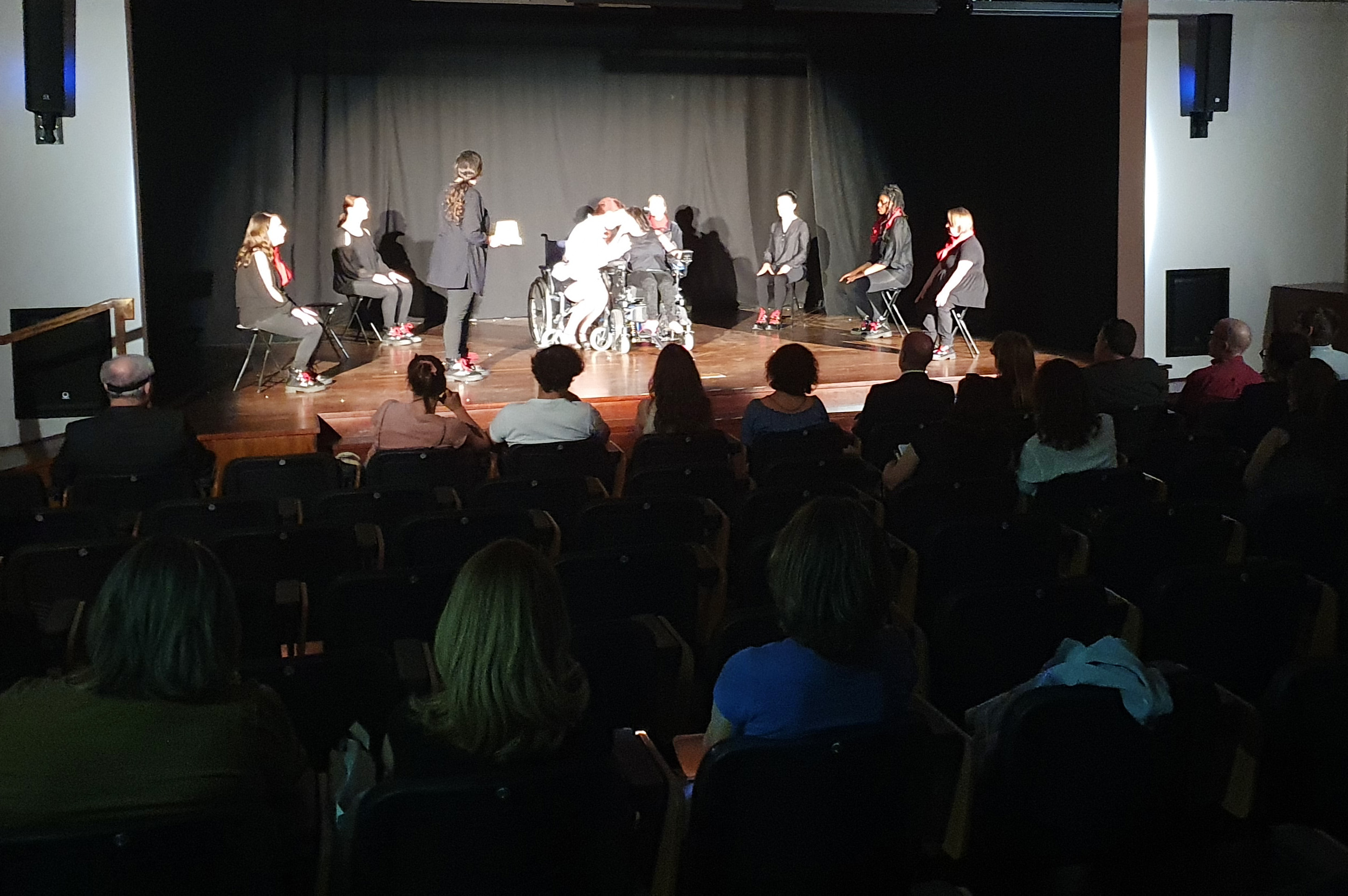 Fotografia de sala de espetáculos, pouco iluminada, com vários atores e atrizes em palco. No público, na penumbra, várias pessoas a assistir.