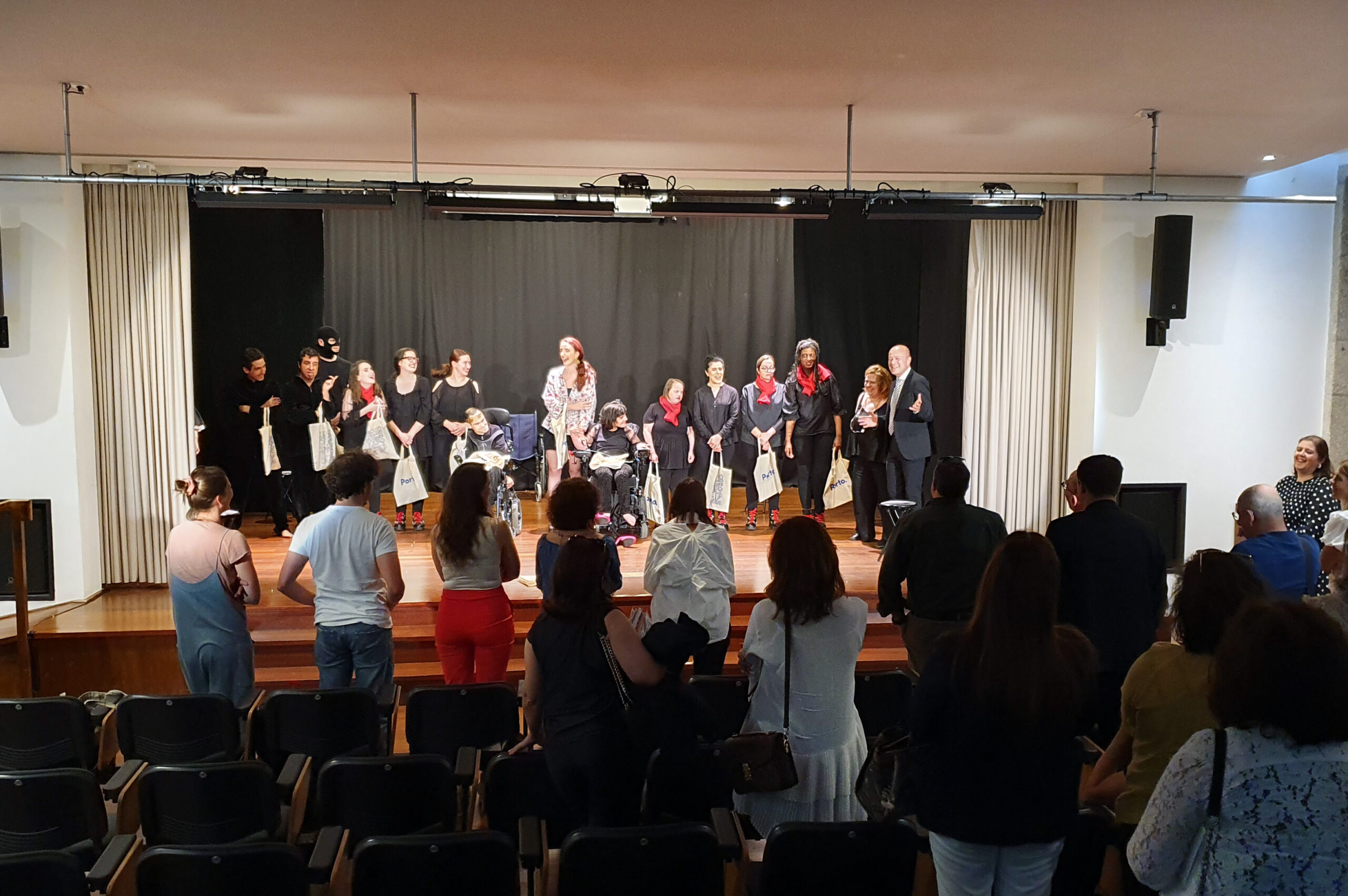 Pessoas de pé a aplaudir atores e atrizes que em palco agradecem. Também no palco estão o Vereador da Câmara do Porto, Fernando Paulo, e a encenadora do grupo de teatro da APPC, Mónica Cunha.