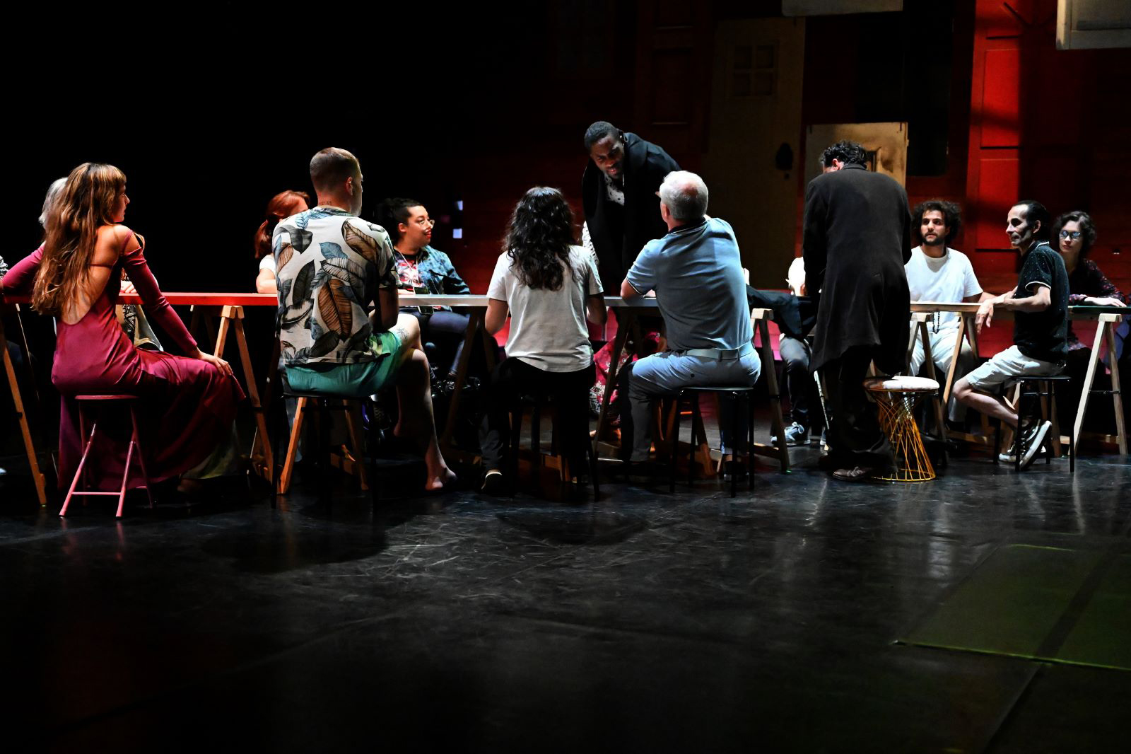 Grupo de atores e atrizes em palco, todos à volta de uma mesa enorme. Excetuando um ator de pé e outro em cima da mesa, de joelhos, todos os restantes es tão sentados.