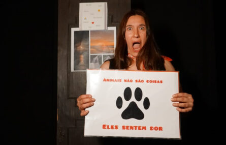 Atriz com ar de surpresa segura cartaz com a frase “Animais não são coisas, eles sentem dor”. Atrás vê-se uma porta em madeira com algumas imagens coladas e uma folha com palavras impercetíveis.