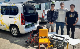 Três sorridentes jovens, lado a lado, junto de alguns dos produtos de apoio oferecidos à Associação do Porto de Paralisia Cerebral. Fotografia de exterior, em zona de estacionamento, estando ao lado dos jovens uma viatura do Colégio Internato dos Carvalhos e sendo ainda visíveis outras viaturas de serviço da Associação do Porto de Paralisia Cerebral