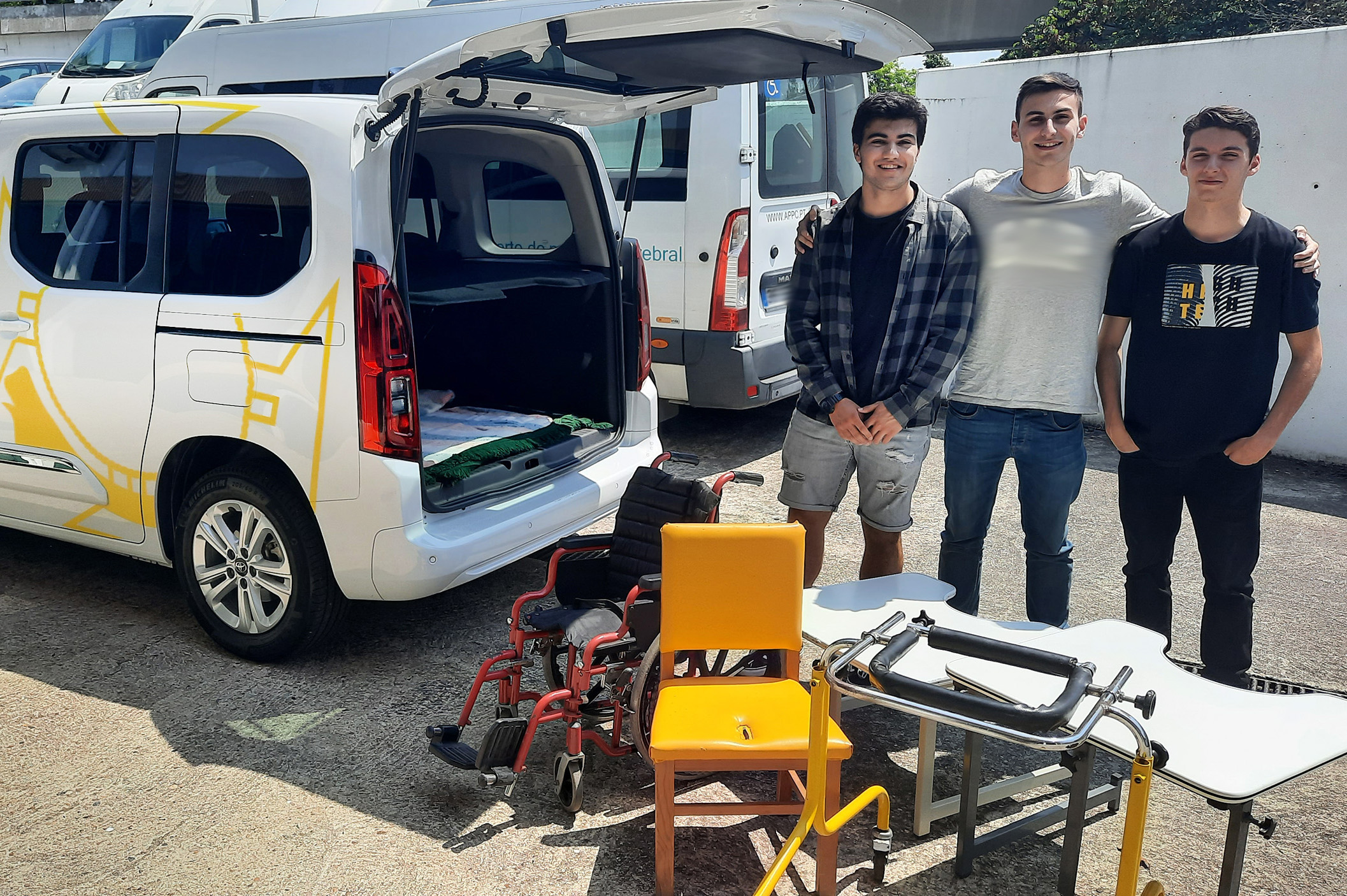 Três sorridentes jovens, lado a lado, junto de alguns dos produtos de apoio oferecidos à Associação do Porto de Paralisia Cerebral. Fotografia de exterior, em zona de estacionamento, estando ao lado dos jovens uma viatura do Colégio Internato dos Carvalhos e sendo ainda visíveis outras viaturas de serviço da Associação do Porto de Paralisia Cerebral
