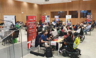 Imagem de espaço aberto em auditório. Inúmeras mesas, cadeiras e pessoas sentadas (algumas em cadeiras de rodas). Estão a decorrer conversas e entrevistas. Alguns cartazes, dispersos, com informação e publicidade às empresas presentes.
