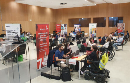 Imagem de espaço aberto em auditório. Inúmeras mesas, cadeiras e pessoas sentadas (algumas em cadeiras de rodas). Estão a decorrer conversas e entrevistas. Alguns cartazes, dispersos, com informação e publicidade às empresas presentes.