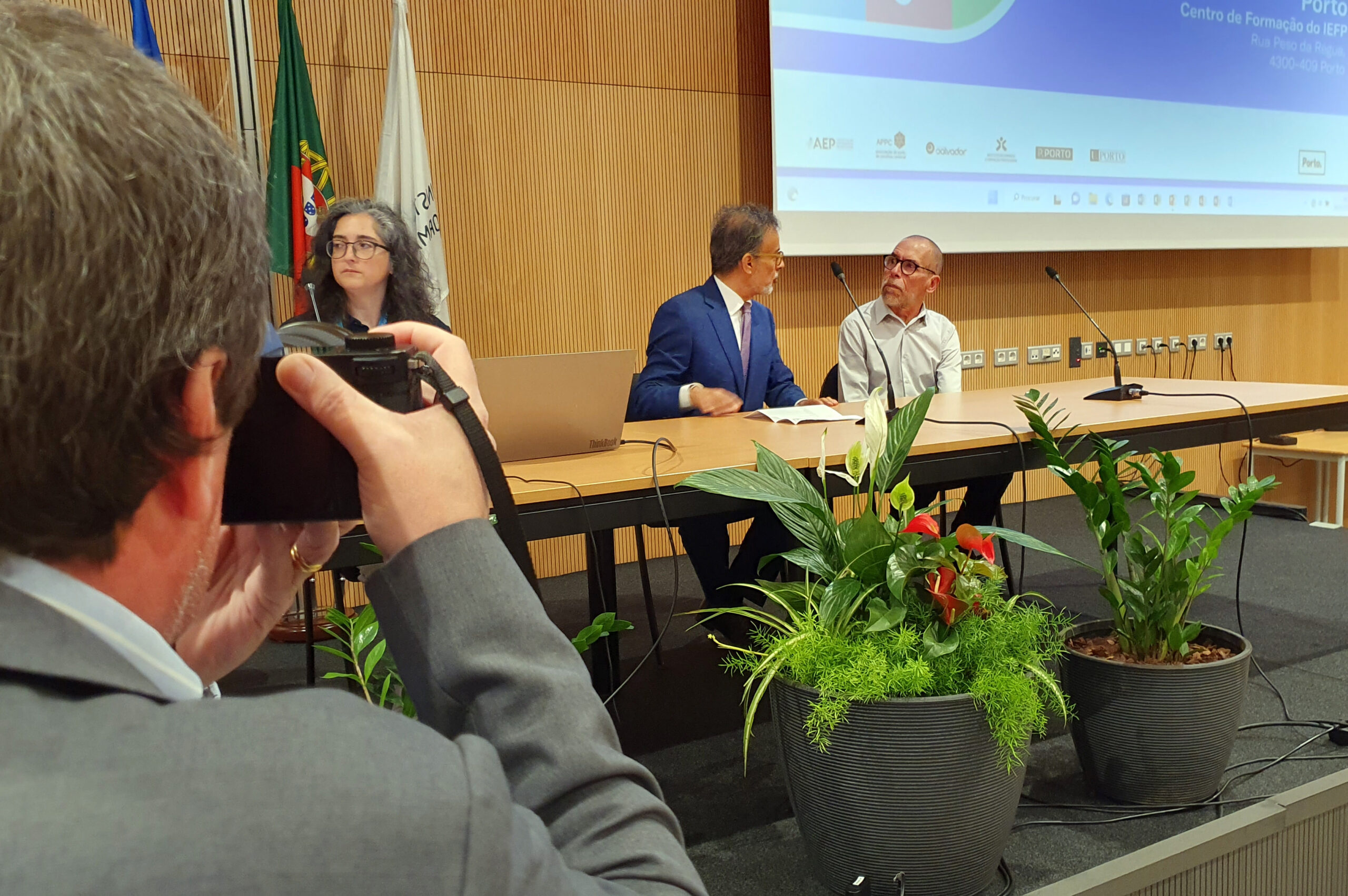 Imagem de espaço aberto em auditório. Inúmeras mesas, cadeiras e pessoas sentadas (algumas em cadeiras de rodas). Estão a decorrer conversas e entrevistas. Alguns cartazes, dispersos, com informação e publicidade às empresas presentes. Ao fundo o palco do auditório e algumas pessoas a prepararem-se para a sessão de abertura.
