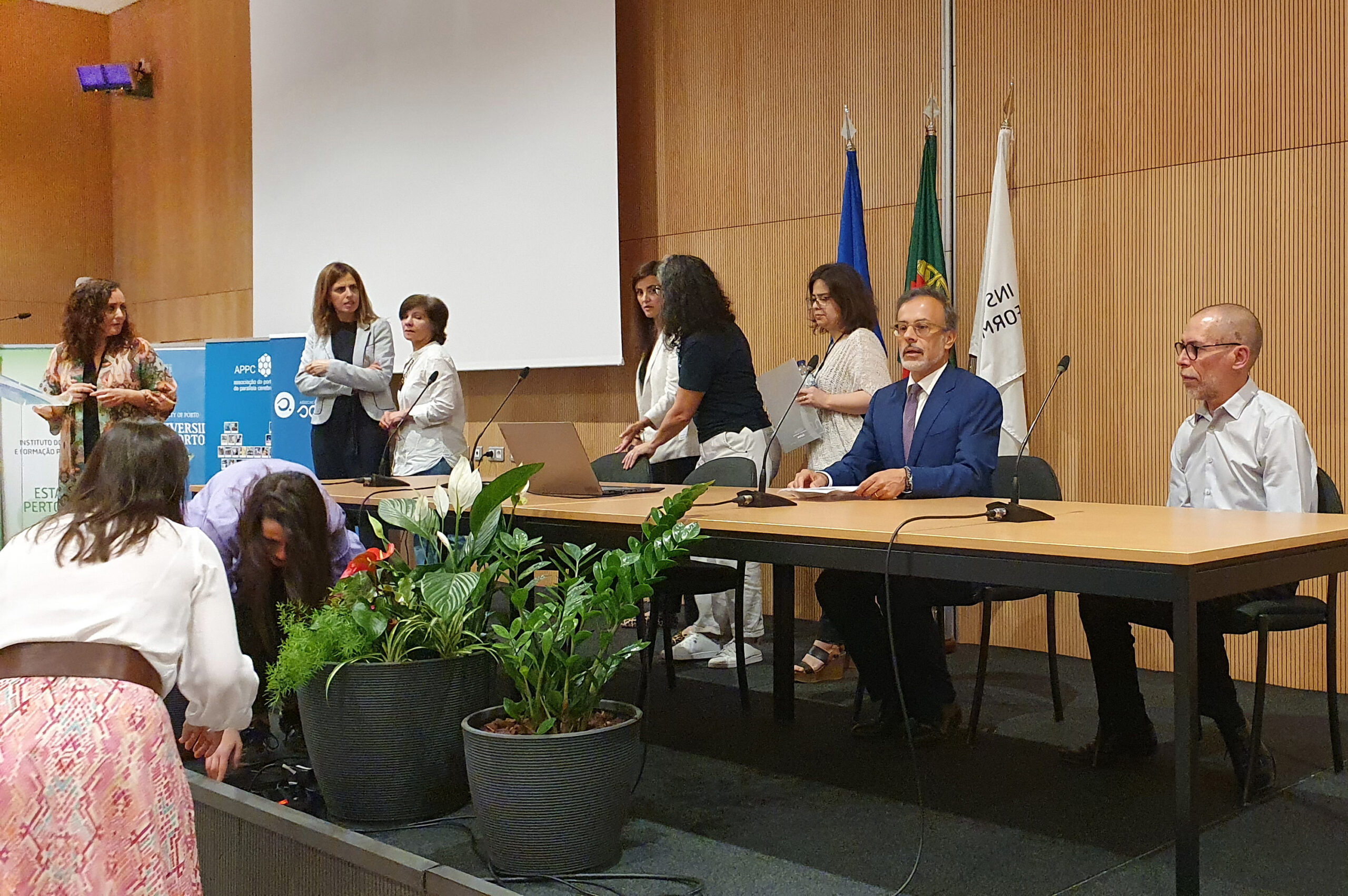 Imagem da mesa de abertura da sessão (ainda com pessoas a sentarem-se). À mesa estão já sentadas duas pessoas. Em primeiro plano dois vasos com plantas.