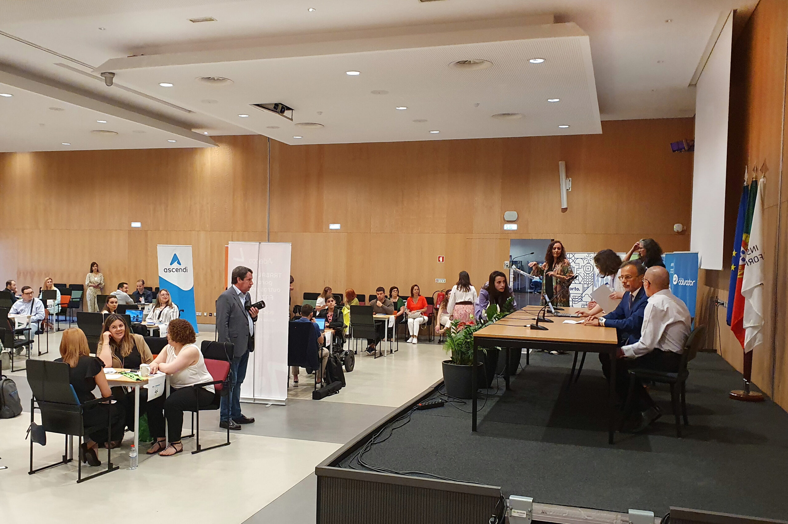 Plano geral com a mesa de abertura da sessão e aspeto parcial do auditório. Estão já a decorrer conversas e entrevistas. Alguns cartazes, dispersos, com informação e publicidade às empresas presentes.