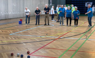 Fotografia de interior de pavilhão desportivo. O chão, em madeira, tem algumas marcações coloridas. Em primeiro cinco bolas de boccia pousadas no chão. Mais ao fundo estão sete jogadores, de duas equipas. No fundo da fotografia estão outras pessoas a conversar e assistir.
