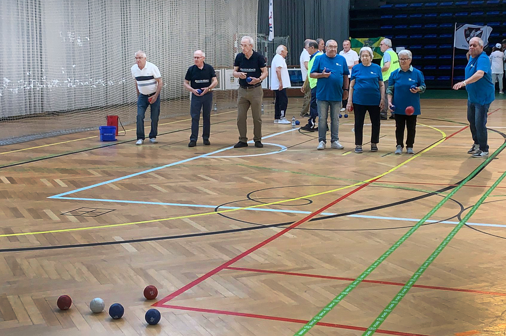 Fotografia de interior de pavilhão desportivo. O chão, em madeira, tem algumas marcações coloridas. Em primeiro cinco bolas de boccia pousadas no chão. Mais ao fundo estão sete jogadores, de duas equipas. No fundo da fotografia estão outras pessoas a conversar e assistir.