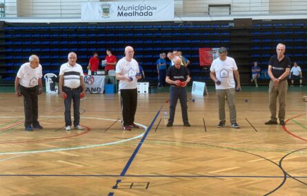 Fotografia de interior de pavilhão desportivo. O chão, em madeira, tem algumas marcações coloridas. Em primeiro plano, alinhados, vários praticantes de boccia. Ao fundo vê-se a bancada, a mesa e apoio e algumas outras pessoas. Pendurado na bancada está cartaz com indicação Município da Mealhada.