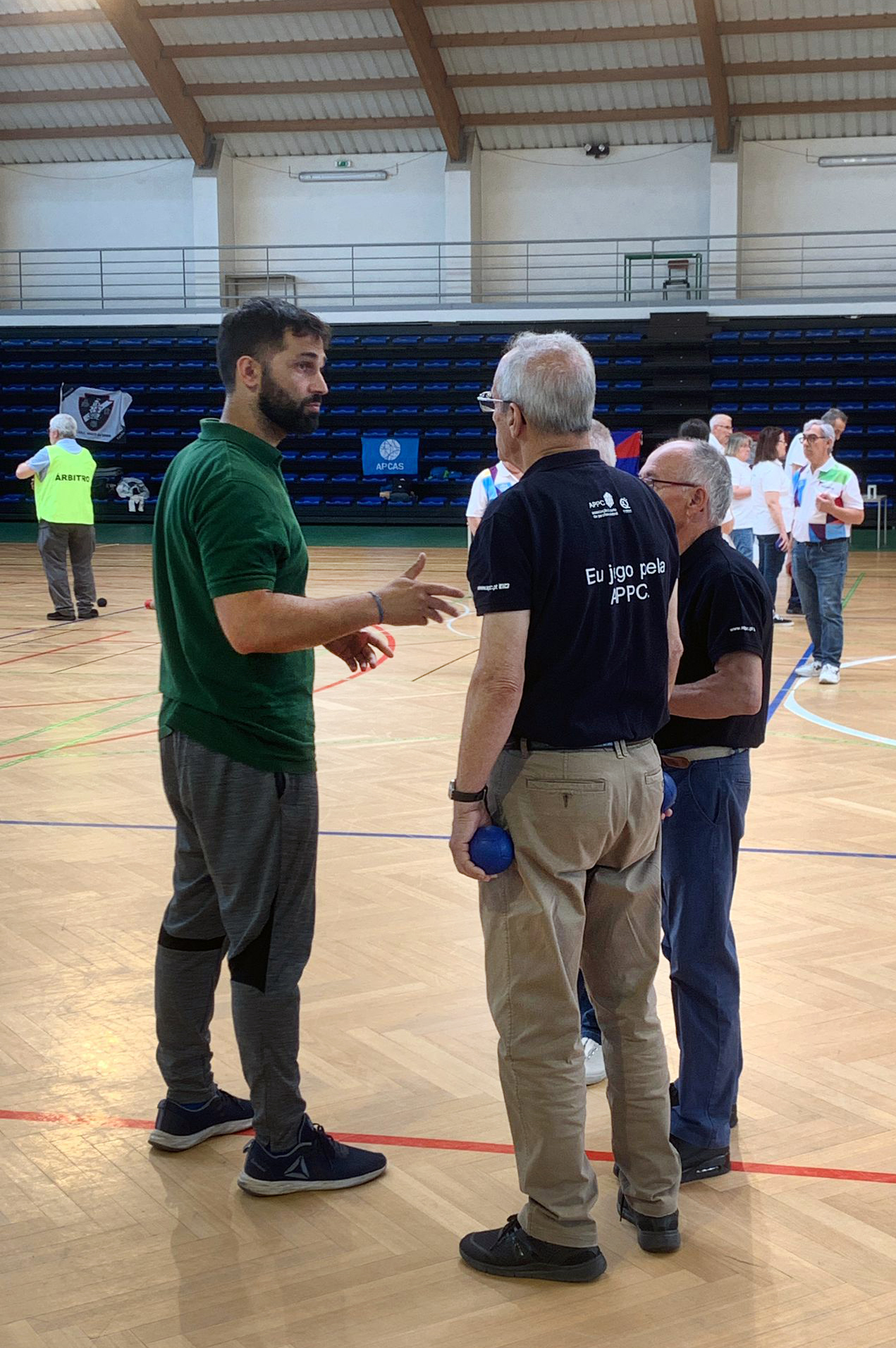 Fotografia de interior de pavilhão desportivo com grupo de três pessoas a conversar. A camisola de uma dessas pessoas, cor azul escura, tem nas costas a expressão “Eu jogo pela APPC”. Ao fundo da imagem estão várias outras pessoas a dialogar e a jogar boccia.