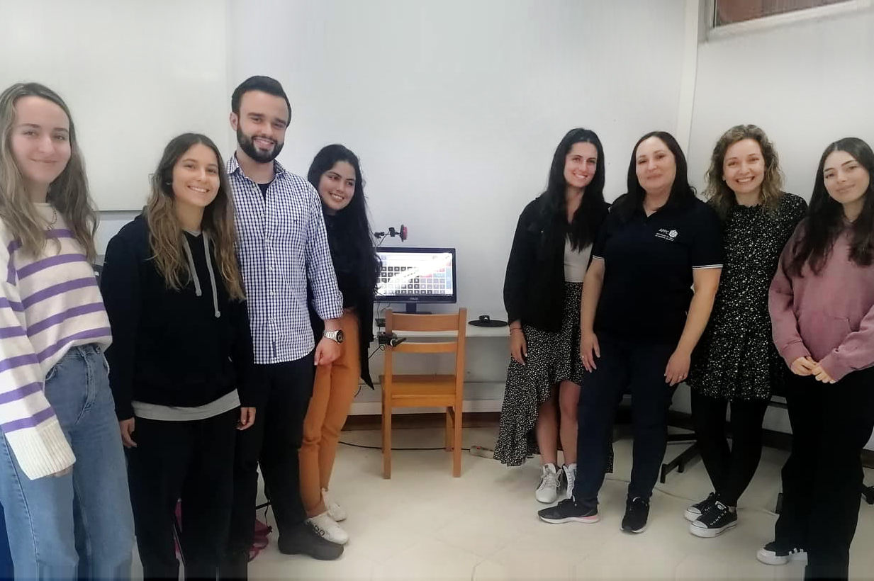 Fotografia de grupo de oito pessoas em espaço interior do Centro de Reabilitação da APPC. Além das pessoas, ao centro, vê-se uma cadeira de madeira e um monitor de computador.