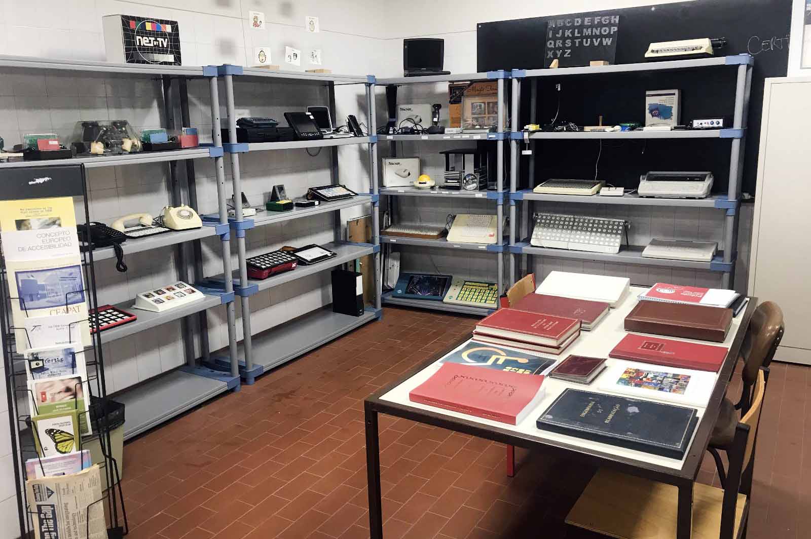 Em primeiro plano uma mesa com vários livros e manuais pousados. Ao fundo quatro conjuntos de estantes com inúmeras tecnologias de apoio.