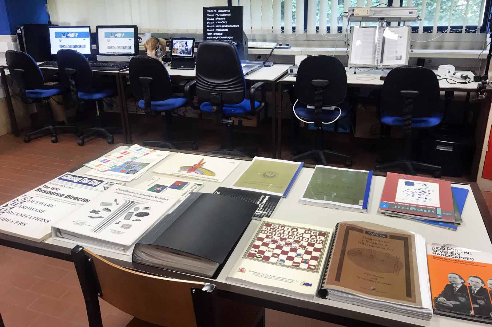 Em primeiro plano uma mesa com diversos manuais e livros técnicos. Mais ao fundo, junto de seis cadeiras, estão três secretárias com diversas tecnologias de apoio.