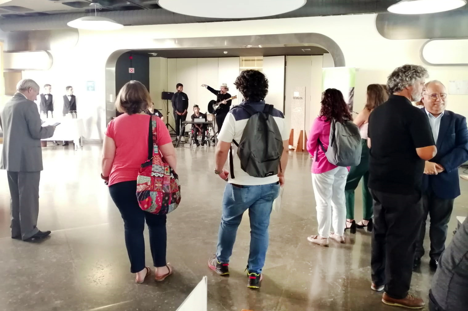 Ao fundo da fotografia está a banda appSound em palco improvisado junto da entrada de um auditório. Em primeiro plano, de costas, algumas pessoas assistem à atuação e outras conversam.