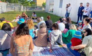 Fotografia de várias pessoas sentadas em cima da relva em jardim exterior. A maioria encontra-se de costas. Mais à direita estão três adultos a assistir de pé (um deles com uma criança ao colo). Todos os rostos das crianças foram digitalmente desfocados.