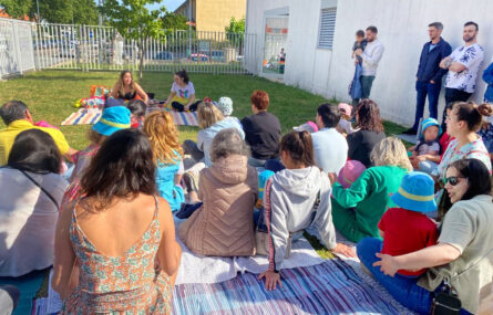 Fotografia de várias pessoas sentadas em cima da relva em jardim exterior. A maioria encontra-se de costas. Mais à direita estão três adultos a assistir de pé (um deles com uma criança ao colo). Todos os rostos das crianças foram digitalmente desfocados.