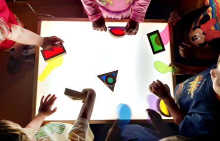 Fotografia em plano superior de várias crianças a brincarem com peças numa mesa de luz. Em cima da mesa estão peças de diferentes formatos e cores.
