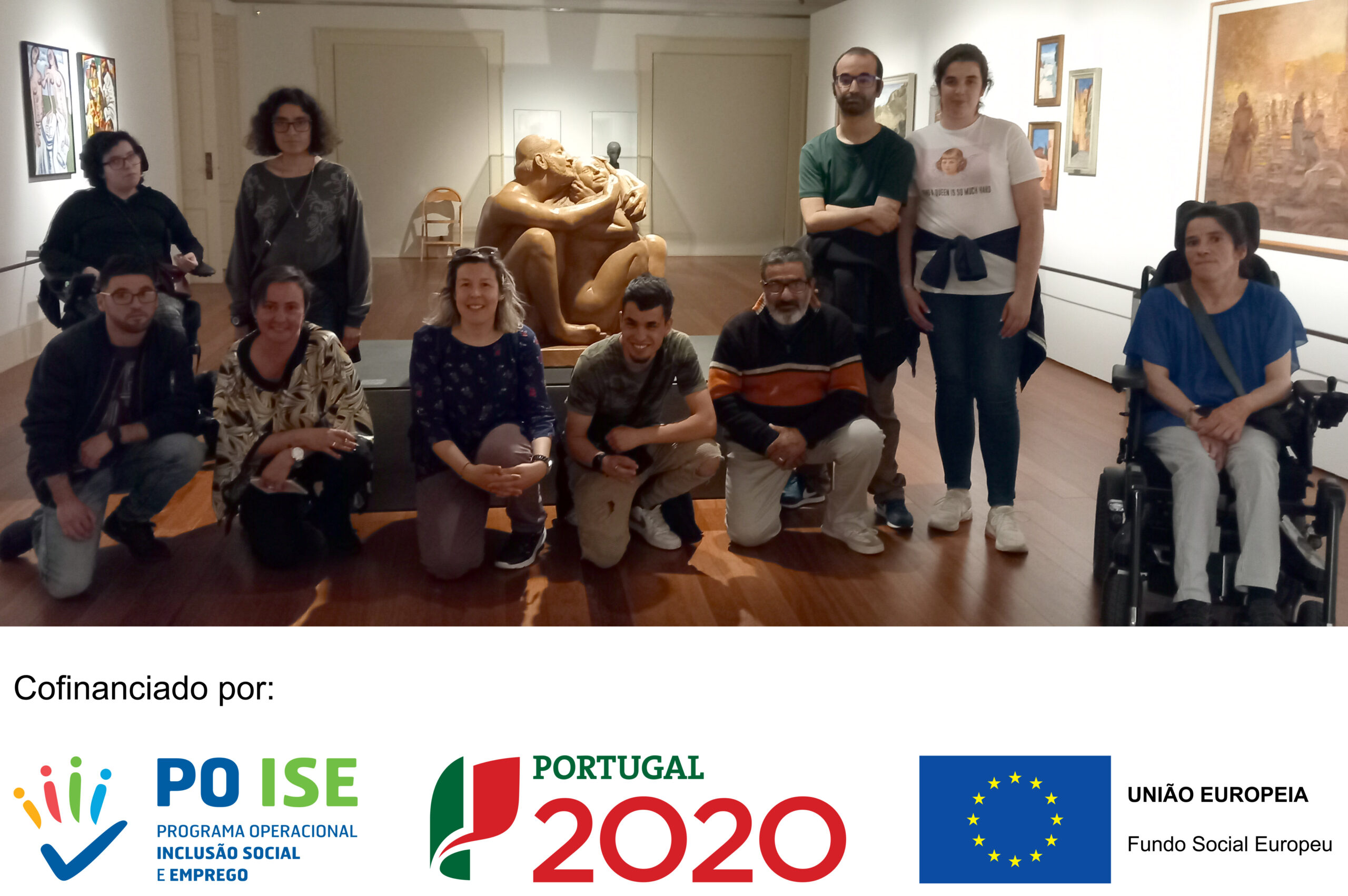 Fotografia de espaço interior de exposições. Nas paredes estão colocados alguns quadros e pinturas. Ao centro, atrás, vê-se uma escultura de duas pessoas a abraçarem-se. Em destaque surgem 10 pessoas, do Curso Assistente Familiar e Apoio à Comunidade. O rodapé da fotografia tem, em fundo branco, logótipos das entidades que cofinanciam o projeto: Programa Operacional Inclusão Social e Emprego; Portugal 2020; e União Europeia – Fundo Social Europeu.