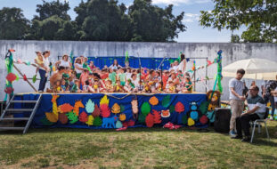 Imagem de um palco (instalado em jardim) com perto de meia centena de crianças e adultos. O palco tem na frente imagens e motivos alusivos ao tema da festa – nomeadamente alguns animais e plantas da selva. Na zona lateral do palco está o Presidente da Direção da APPC, Abílio Cunha, a fazer uma intervenção (enquanto outra pessoa lhe segura o microfone). Ao fundo vê-se um muro e atrás desse muro várias árvores. Os rostos das crianças encontram-se todos desfocados.