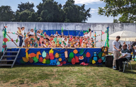 Imagem de um palco (instalado em jardim) com perto de meia centena de crianças e adultos. O palco tem na frente imagens e motivos alusivos ao tema da festa – nomeadamente alguns animais e plantas da selva. Na zona lateral do palco está o Presidente da Direção da APPC, Abílio Cunha, a fazer uma intervenção (enquanto outra pessoa lhe segura o microfone). Ao fundo vê-se um muro e atrás desse muro várias árvores. Os rostos das crianças encontram-se todos desfocados.