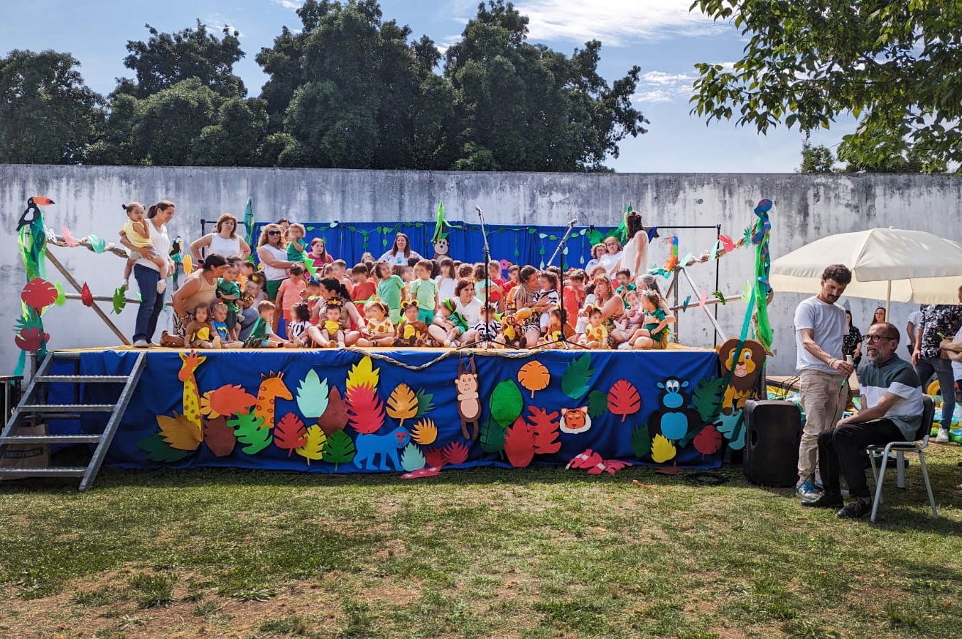 Imagem de um palco (instalado em jardim) com perto de meia centena de crianças e adultos. O palco tem na frente imagens e motivos alusivos ao tema da festa – nomeadamente alguns animais e plantas da selva. Na zona lateral do palco está o Presidente da Direção da APPC, Abílio Cunha, a fazer uma intervenção (enquanto outra pessoa lhe segura o microfone). Ao fundo vê-se um muro e atrás desse muro várias árvores. Os rostos das crianças encontram-se todos desfocados.