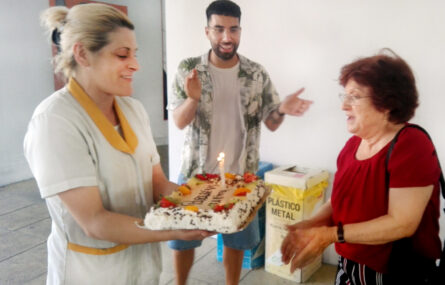 Três pessoas na imagem. À esquerda há uma pessoa que segura um bolo de aniversário com vela acesa. À direita a aniversariante prepara-se para apagar a vela. Ao centro está uma pessoa a bater palmas e a sorrir.