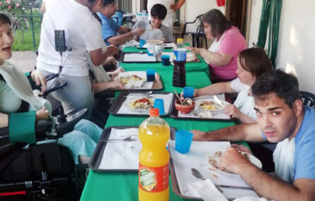 Fotografia de várias pessoas sentadas junto de uma mesa comprida (com comidas e bebidas).