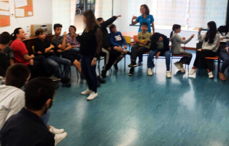 Mais de uma dezena de crianças e jovens a conversar, sentados em cadeiras, em semí-círculo, em sala interior. Estão acompanhados por duas pessoas adultas. Todos os rostos de crianças e jovens estão desfocados.