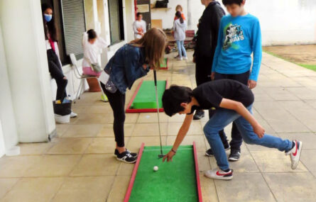 Grupo de crianças a jogar mini-golfe. Enquanto uns jogam, outros assistem. Todos os rostos de crianças e jovens estão desfocados.