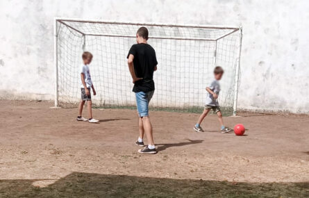 Três crianças com uma bola em jogo de futebol. Ao fundo está a baliza. Todos os rostos de crianças e jovens estão desfocados.