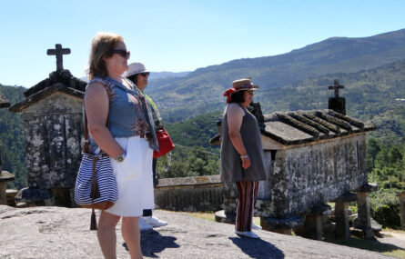 Várias pessoas, de perfil, olha para a paisagem. Ao fundo dois dos espigueiros do Soajo e, ainda mais distante, montes e céu azul.