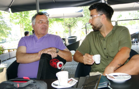 Dois homens sentados a conversar em esplanada. Um deles segura uma chávena de café.