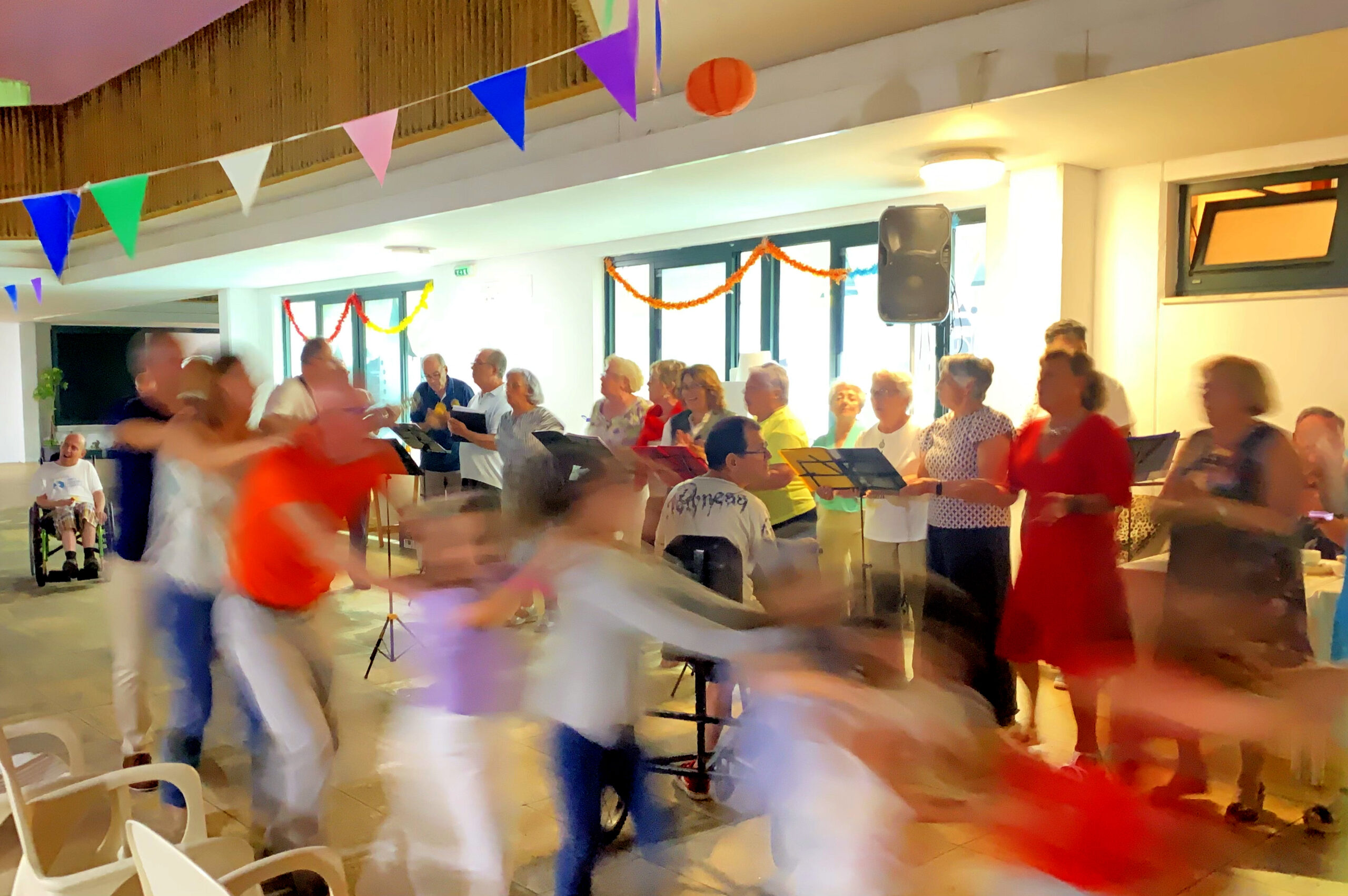 Ao fundo da imagem um grupo de pessoas a cantar. À frente do grupo estão várias pessoas, em fila, a dançar. A fila de pessoas está completamente desfocada (porque a imagem foi feita com captura lenta e, assim, as pessoas ficaram com aspeto “arrastado”). À esquerda da imagem está, sorridente, uma pessoa sentada em cadeira de rodas.
